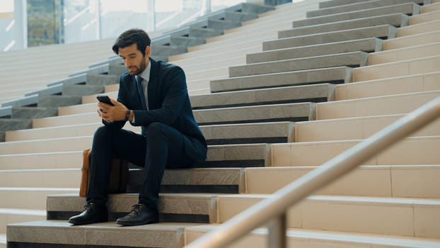 Smart project manager looking at mobile phone while sitting at stairs. Attractive caucasian business man working by using phone. Professional investor using phone to plan business strategy. Exultant.