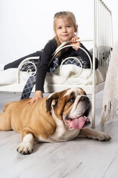 In the girl's bedroom there is an English bulldog next to her bed and the child is lying on the bed and accosts the dog. A breed with a brown coat with white patches.