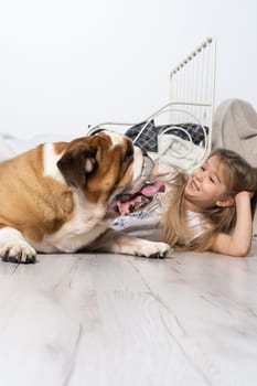 An old and fat dog and a tiny and young girl sit on the floor by the bed in the bedroom. A breed with a brown coat with white patches.