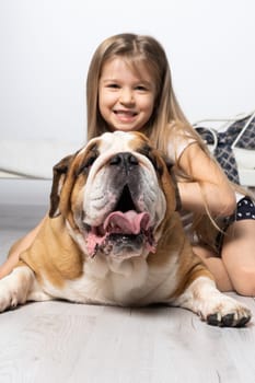 An old and fat dog and a tiny and young girl sit on the floor by the bed in the bedroom. A breed with a brown coat with white patches.