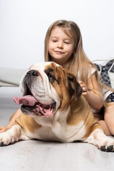 An old and fat dog and a tiny and young girl sit on the floor by the bed in the bedroom. A breed with a brown coat with white patches.