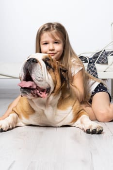 An old and fat dog and a tiny and young girl sit on the floor by the bed in the bedroom. A breed with a brown coat with white patches.
