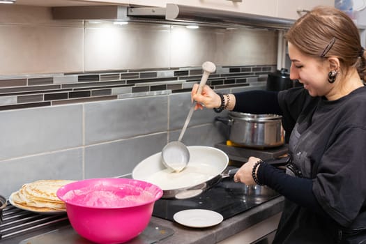 In the kitchen, a girl is standing by an induction cooker, pouring the ready-made pancake batter into a hot pan. Homemade pancakes from a woman's hand.