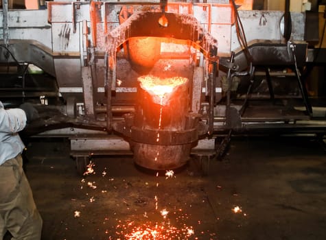 Molten metal being poured from a ladle into a container, sparks flying in an industrial setting. Steel mill, factory, or foundry. Industrial metal production, casting, or pouring concept