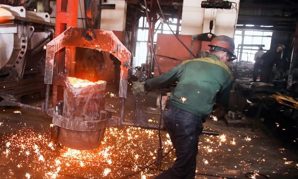 Steel mill worker wearing safety gear and a hard hat carefully pours glowing molten metal into a mold, creating sparks as industrial machinery hums around them in a busy factory setting.