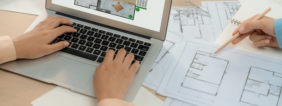 Eco house blueprint displayed on a laptop. Professional architect hand draws blueprint on meeting table with architectural document scatter around. Focus on hand. Closeup. Delineation.