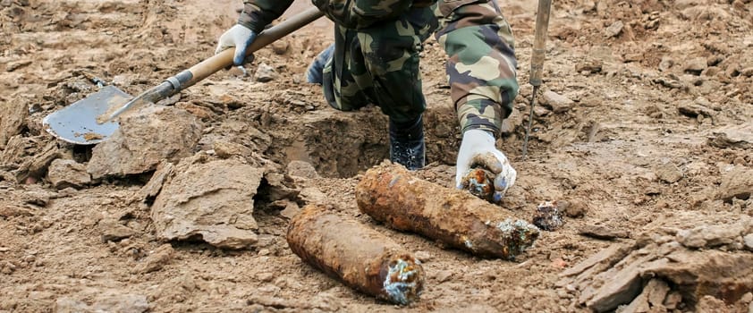 Soldier in protective gear carefully removes live mortar shell from ground during demining operation. Soldier uses shovel to dig around shell. Image shows barren landscape in background.