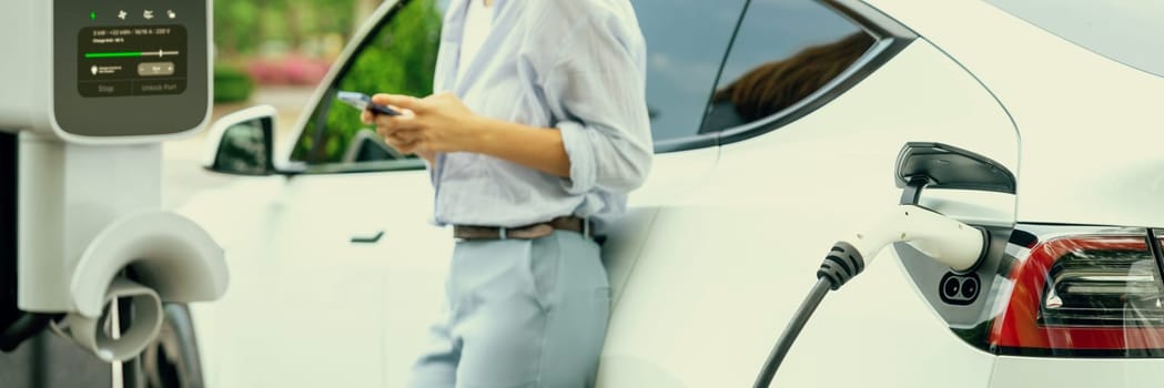 Young woman using smartphone online banking application to pay for electric car battery charging from EV charging station during vacation road trip at national park or summer forest. Panorama Exalt