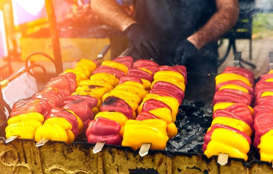 Red and green peppers grilling on barbecue, smoke rising. Person in apron flipping with tongs. Summertime barbecue vibes, roasting peppers aroma, street food, culinary expertise.