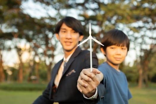 Asian boy and businessman holding wind turbine model together as Earth day concept as corporate social responsible to make greener with eco alternative energy for sustainable future generation. Gyre