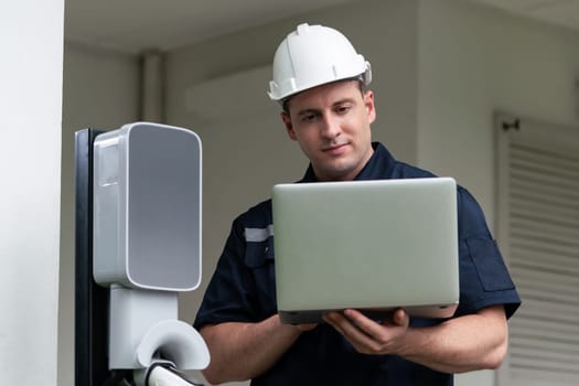 Qualified technician working on home EV charging station installation, making troubleshooting and configuration setup on charging system with laptop for EV at home. Synchronos