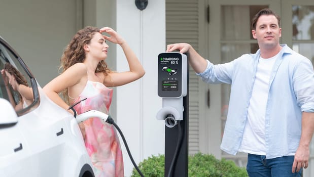 Happy and lovely couple with eco-friendly conscious recharging electric vehicle from EV home charging station. EV car technology utilized for residential home to future sustainability. Synchronos