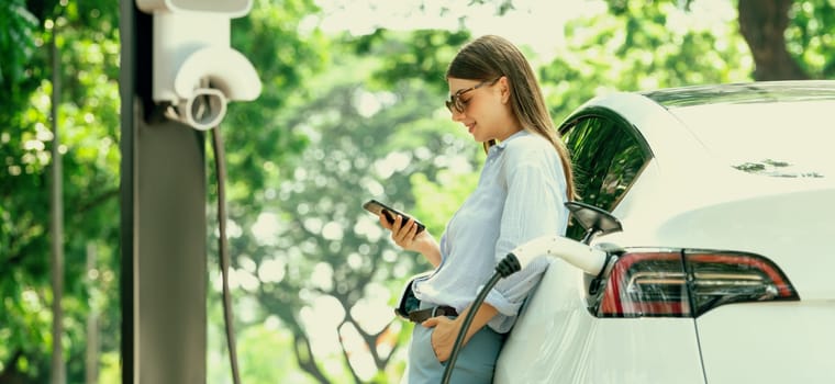 Young woman using smartphone online banking application to pay for electric car battery charging from EV charging station during vacation road trip at national park or summer forest. Panorama Exalt