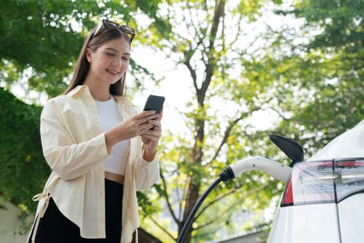 Young woman use smartphone to pay for electricity at public EV car charging station green city park. Modern environmental and sustainable urban lifestyle with EV vehicle. Expedient