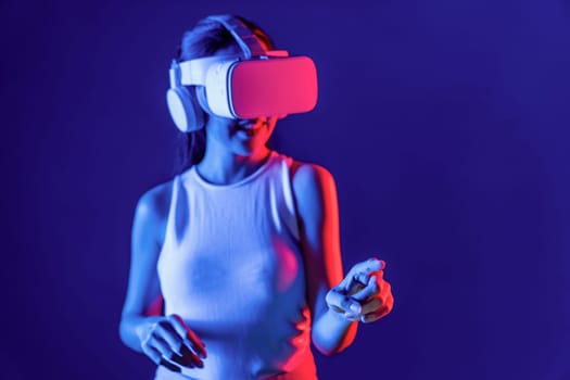 Smart female stand with surrounded by cyberpunk neon light wear VR headset connecting metaverse, futuristic cyberspace community technology. Woman using finger pointing virtual object. Hallucination.