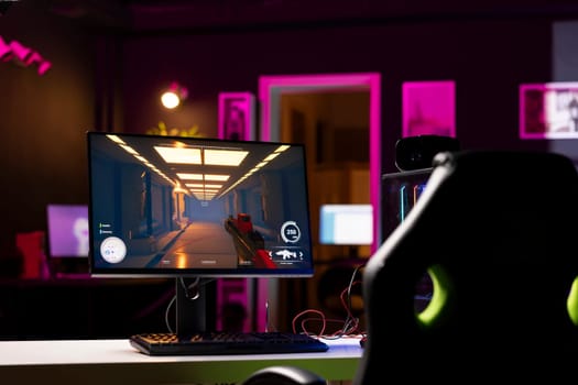 Professional gaming empty room studio with neon lights and RGB lit powerful computer, first person shooter game left open on screen. FPS videogame on pc display in apartment late at night