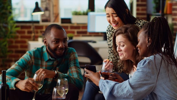 Multiracial friends at apartment party looking at funny videos on cellphone screen. Joyful guests in living room watching internet clips on mobile phone while drinking wine and champagne