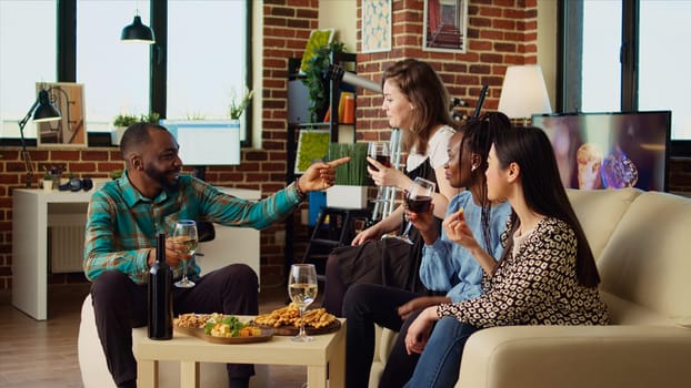 Multiracial group of friends talking with each other in stylish apartment, gathered together to celebrate holiday. Multiethnic colleagues socializing at home, enjoying alcoholic drinks