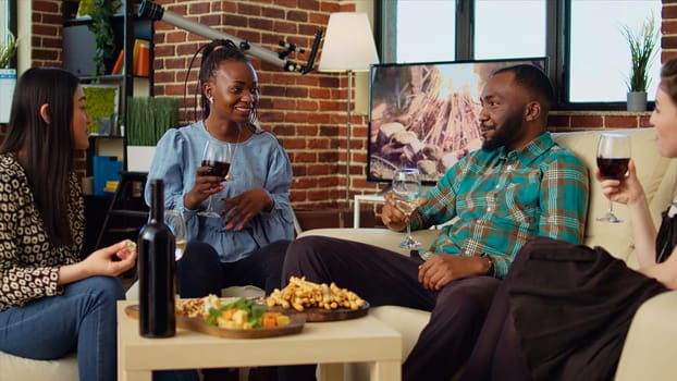 Diverse mates having engaging conversation during apartment party in cozy house. Happy group of people talking in living room, eating snacks and drinking alcoholic beverages