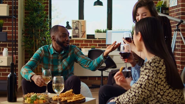 Diverse friends having fun together, laughing and enjoying alcohol drinks and snacks in cozy apartment. BIPOC group of people discussing at home, gathered together for birthday party