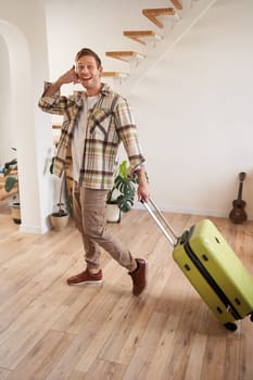 Full length photo of happy guy with suitcase, walking in apartment, shows phone call gesture. Copy space