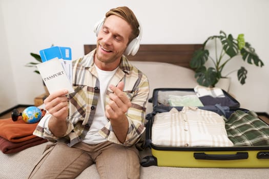 Happy tourist, young man shows passport and tickets, goes on holiday, sits with packed suitcase on bed, plans summer vacation.