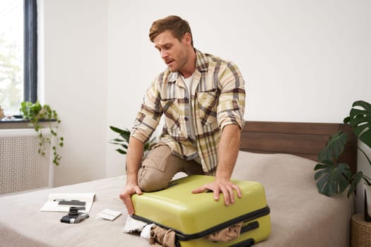Image of young man, tourist struggling to close his suitcase full of clothes, pushing on bag, trying to press items inside luggage to shut the zipper, packing for travelling abroad, going on holiday,