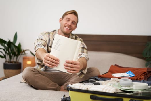 Portrait of smiling, handsome man, packing things in suitcase, showing his written list in notebook, going on holiday. Tourism and lifestyle concept