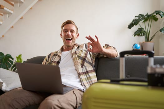 Happy man booking holiday online on laptop, paying for vacation on travel agency website, buying flight tickets in internet, sitting in living room with suitcase.