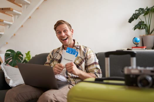 Portrait of happy tourist, man looking excited at laptop screen, holding flight tickets, going on vacation, excited about holiday, sits with suitcase in living room.