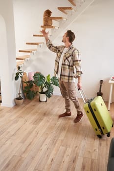 Vertical shot of smiling man with suitcase, playing with a cat, standing and carrying luggage. People concept
