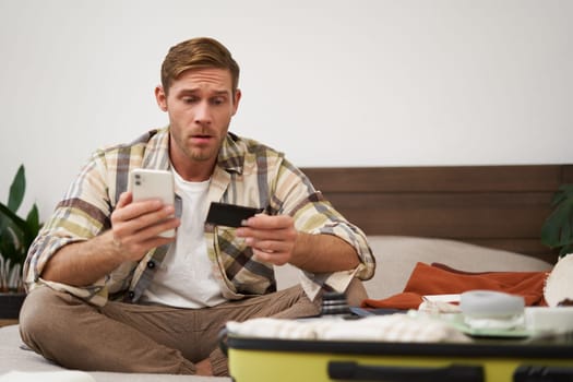 Portrait of young man, tourist paying online, using credit card and mobile phone app, sitting on bed with suitcase, ordering in internet, buying holiday tickets.