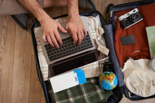 Upper angle view of male hands typing, using laptop on top of the opened suitcase, man packing for holiday, going on vacation, check-in on a flight.