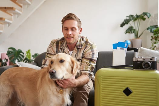 Portrait of young man, traveller, pets his dog, sits with suitcase and tickets, goes on holiday. Copy space
