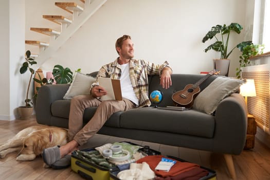 Portrait of young man sitting on sofa with notebook, has dog lying next to him, packing suitcase for summer vacation. Tourism concept