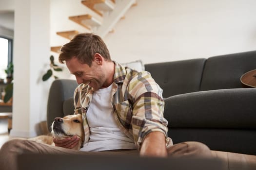 Happy young smiling guy in living room, sitting on floor, hugging his pet, playing with dog. People and animals concept