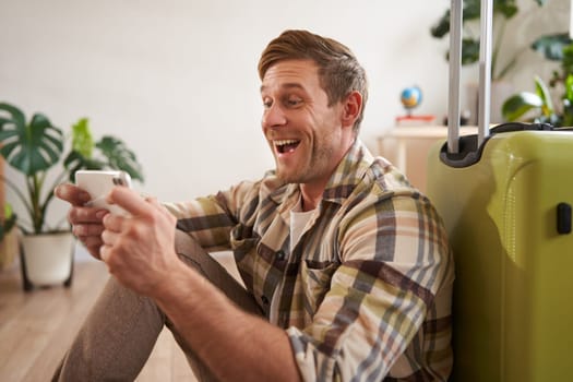 Smiling man with mobile phone, sits near suitcase, laughing while looking at smartphone screen. Travelling concept
