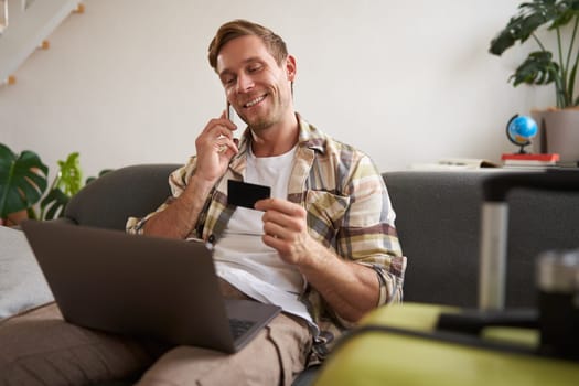 Portrait of smiling guy, tourist booking hotel, confirming purchase over the phone, holding credit card and laptop.