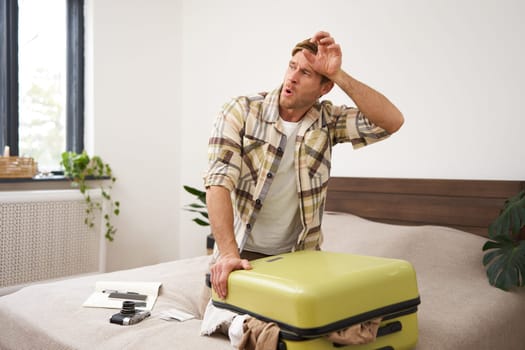 Portrait of young man sweating, shutting his suitcase, trying to close the luggage with lots of clothes, sitting on bed, packing things for summer vacation, going on holiday or business trip.
