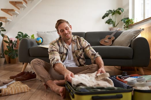 Travelling and tourism concept. Happy young man, sits on floor with luggage opened, packing suitcase for holiday, going on vacation, putting clothes inside a bag.