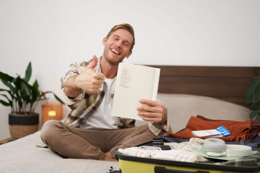 Travelling and tourism. Young man sits on bed, shows his list of items, demonstrates notebook, packs clothes, prepares to go on holiday, goes on vacation, assembles luggage.