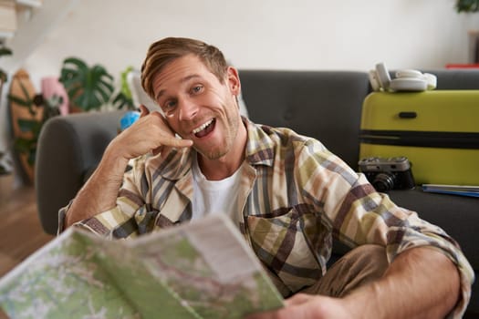 Portrait of handsome smiling young man, tourist shows call me gesture, phone hand, holding travel map, inviting to go on holiday, packed suitcase.