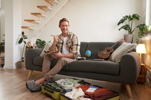Happy man with pen and notebook, checking list of items and packing suitcase for journey, going on holiday, preparing for summer vacation, sitting in living room.