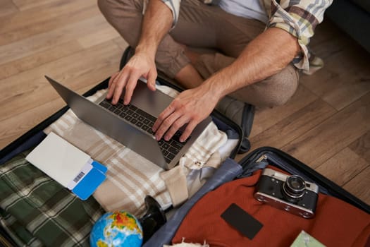Close up shot of man, male hands using laptop on top of opened suitcase, packing for a holiday, going on vacation, prepare camera, two flight tickets for a trip abroad.