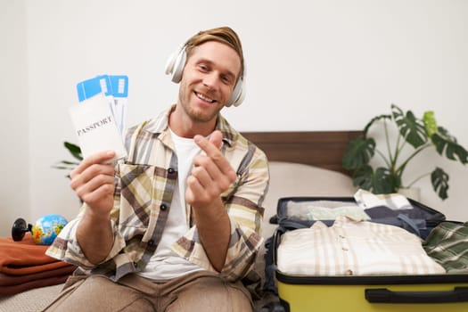Image of handsome young man, tourist shows two plane tickets and passport, travels on holiday, goes on vacation, packs suitcases.