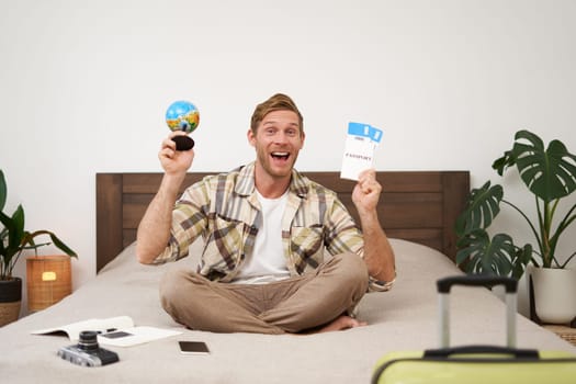 Portrait of cheerful, happy tourist, man sitting with plane tickets and a globe, packed suitcase for travelling around the world, going on vacation, excited about his holiday.