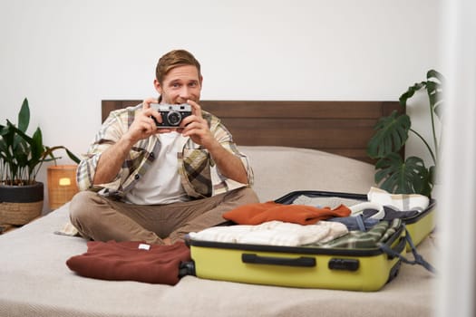 Portrait of tourist, man sits with suitcase on bed, holds camera, packs suitcase with clothes, goes on holiday, prepares for summer vacation, tourist travels abroad.