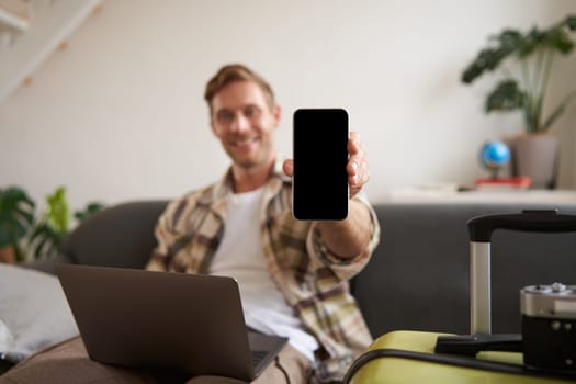 Portrait of handsome young man, showing smartphone screen, recommending mobile app for travelling or booking online, sitting with laptop and suitcase. Copy space