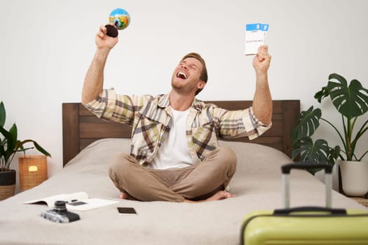 Image of happy smiling man, cheerful tourist sits with globe and plane tickets, goes on holiday, sits with suitcase, excited about his trip, going on holiday. Tourism and travel agency concept
