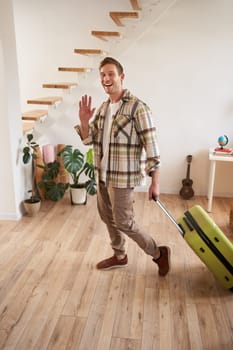 Vertical shot of happy young man with suitcase, checking in the rented apartment, waving hand at camera, walking with luggage indoors.